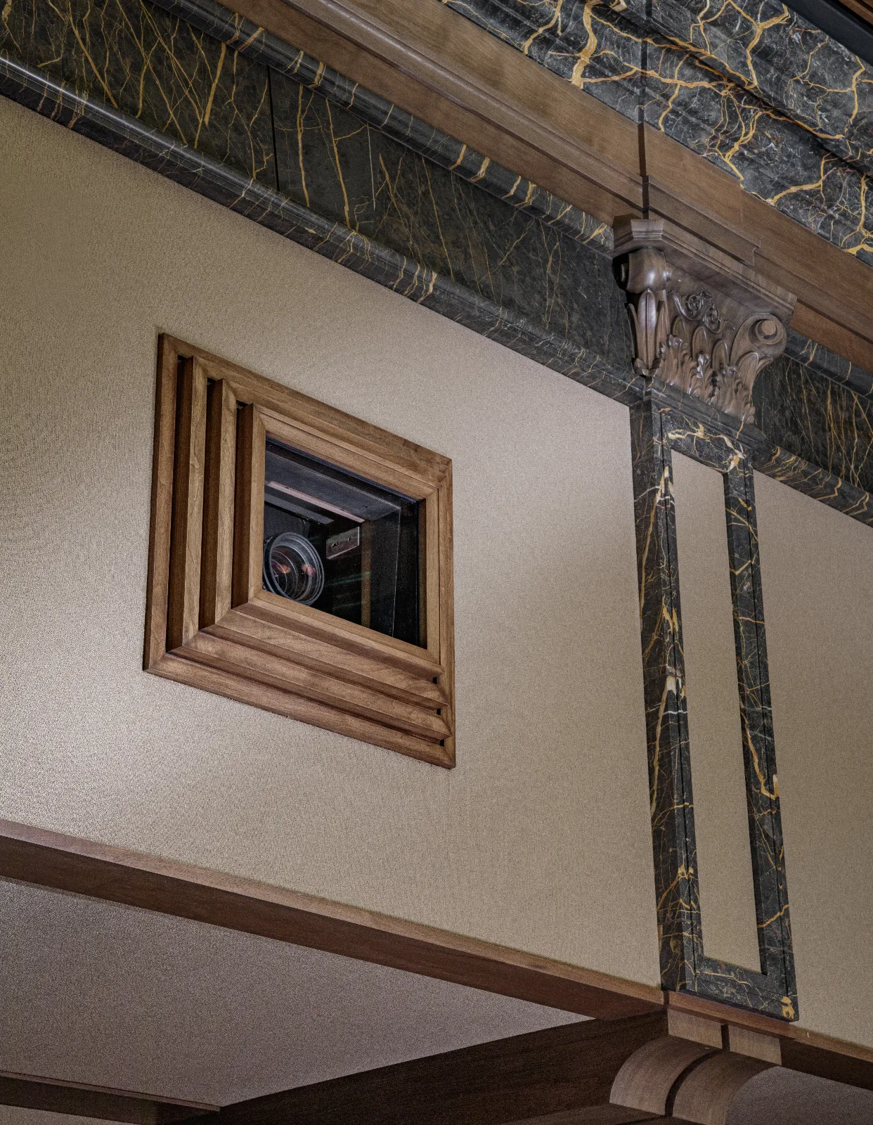 A small wooden framed window containing a projector lens set into a textured wall, with ornate dark marble and wood trim.