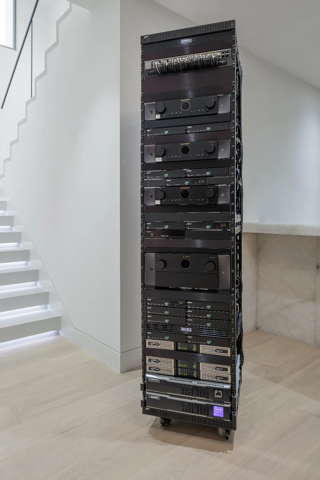 A tall black rack of audio and video equipment on wheels sits on a light wooden floor next to a white modern staircase.