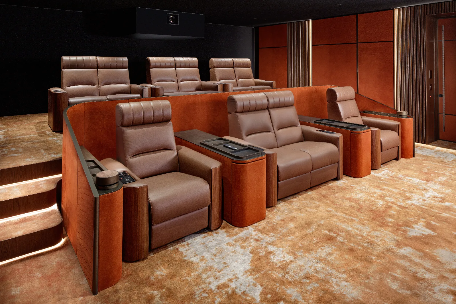 A luxurious home theater with tiered brown leather recliners, orange upholstered walls, and a plush patterned carpet.