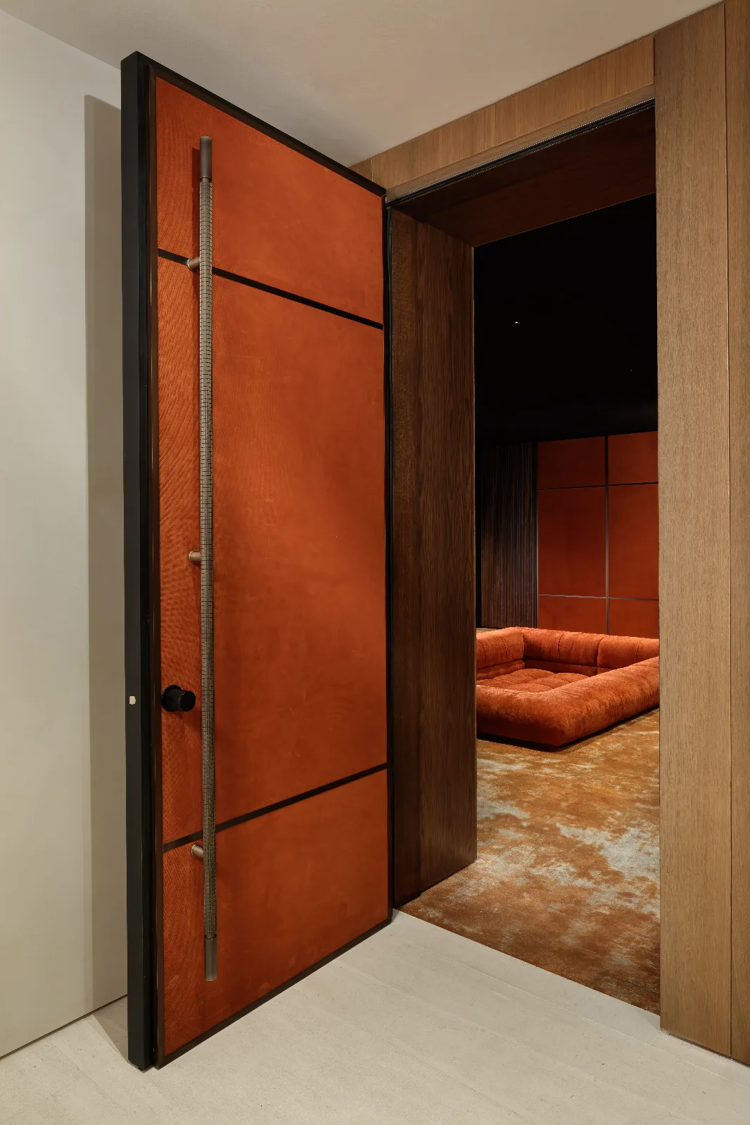 A modern, orange upholstered door with a long metal handle opens into a dark room with a matching sofa and rug.