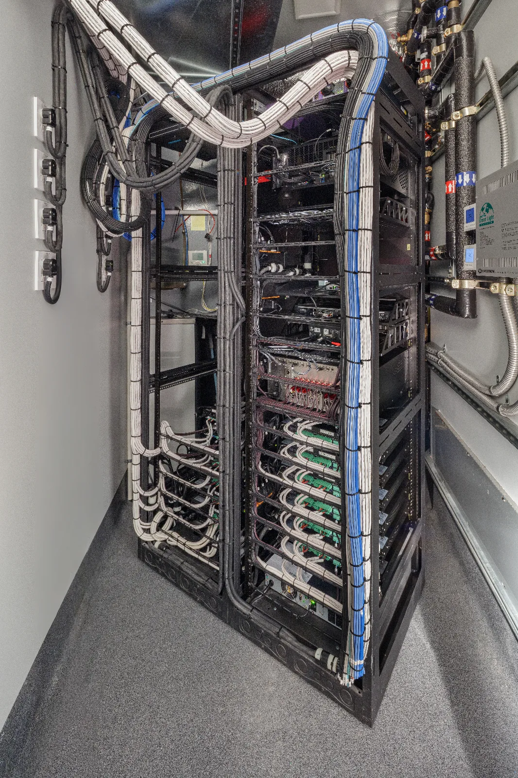 A server rack brimming with neatly bundled white, black, and blue network cables, with power outlets on a white wall.