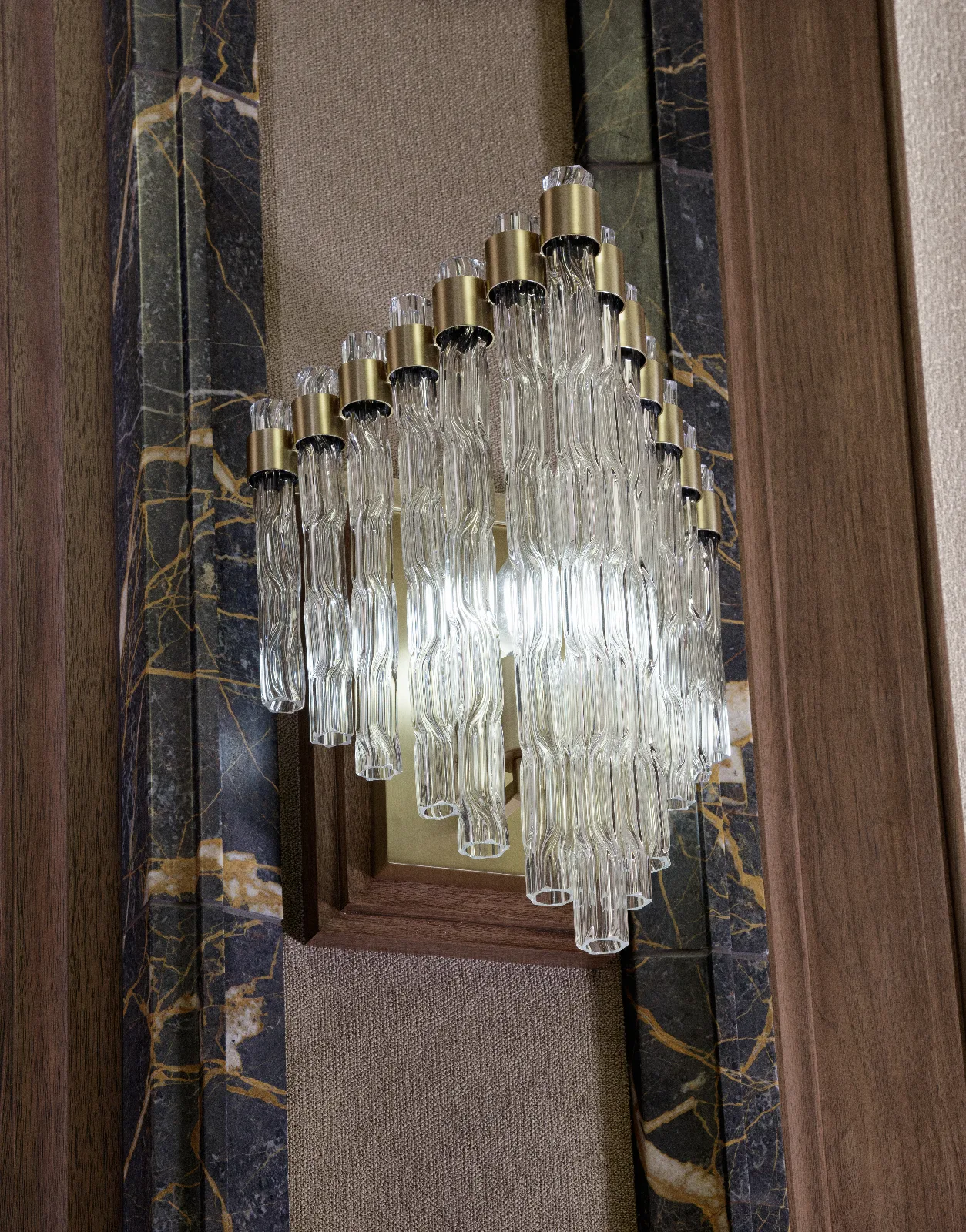 A modern wall sconce features clear textured glass tubes and brass accents, mounted on a textured wall between dark marble and wood panels.