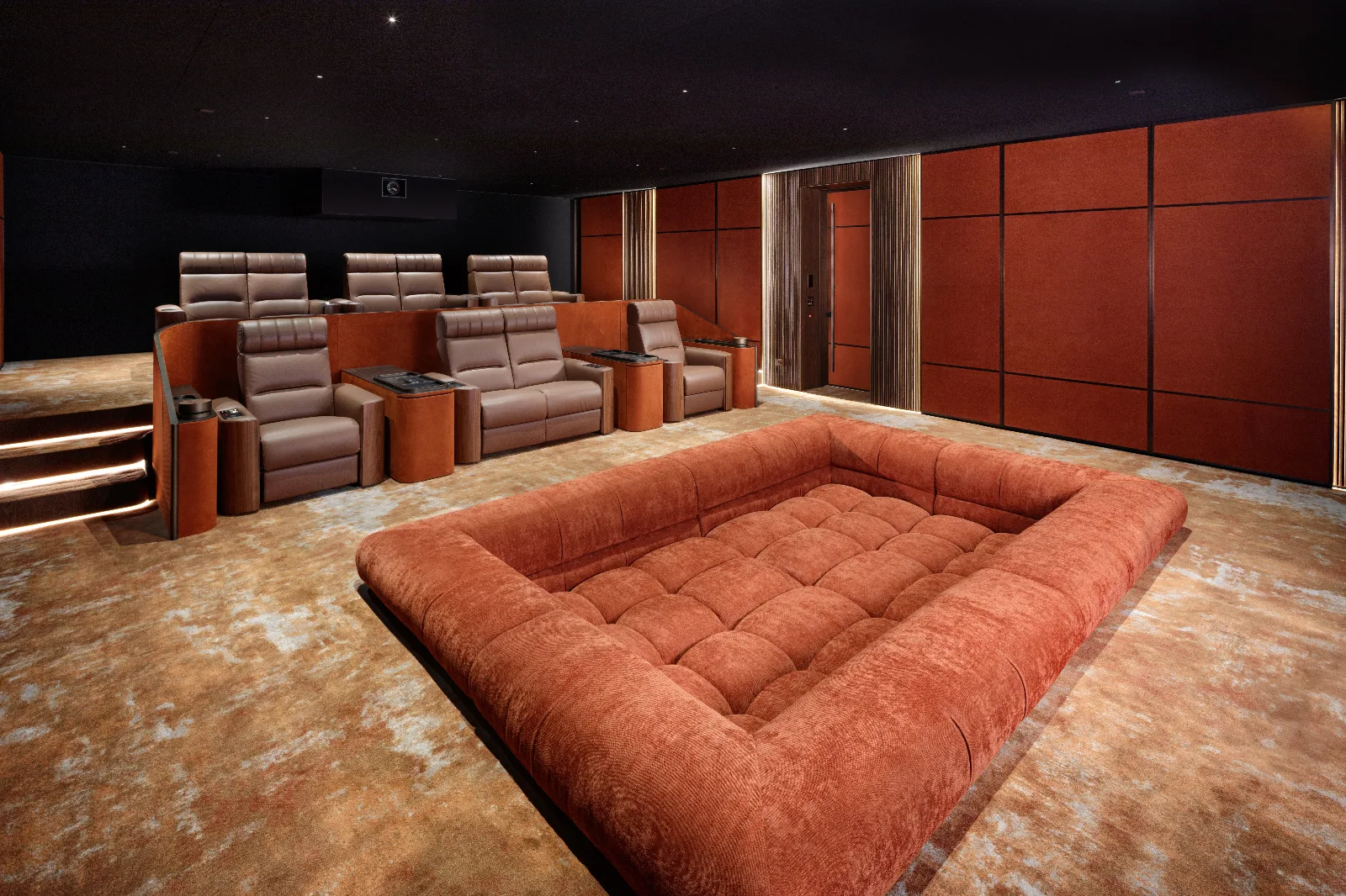 A luxurious home theater features tiered brown recliners, a large orange floor cushion, and sound-absorbing wall panels.