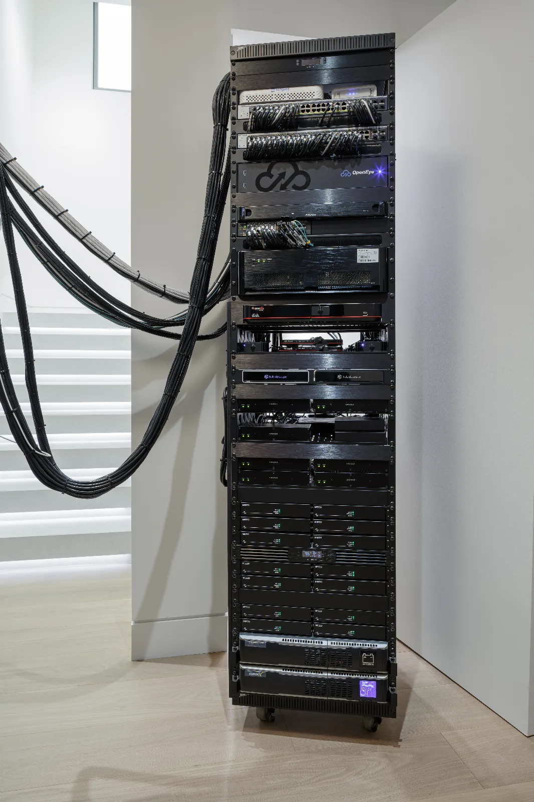 A tall, black server rack filled with electronic components and organized black cable bundles running towards a white staircase.