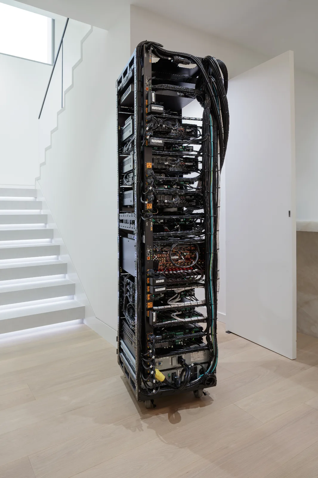 The back of a tall, black server rack filled with dense electronics and tangled black cables, on a light wood floor next to white stairs.