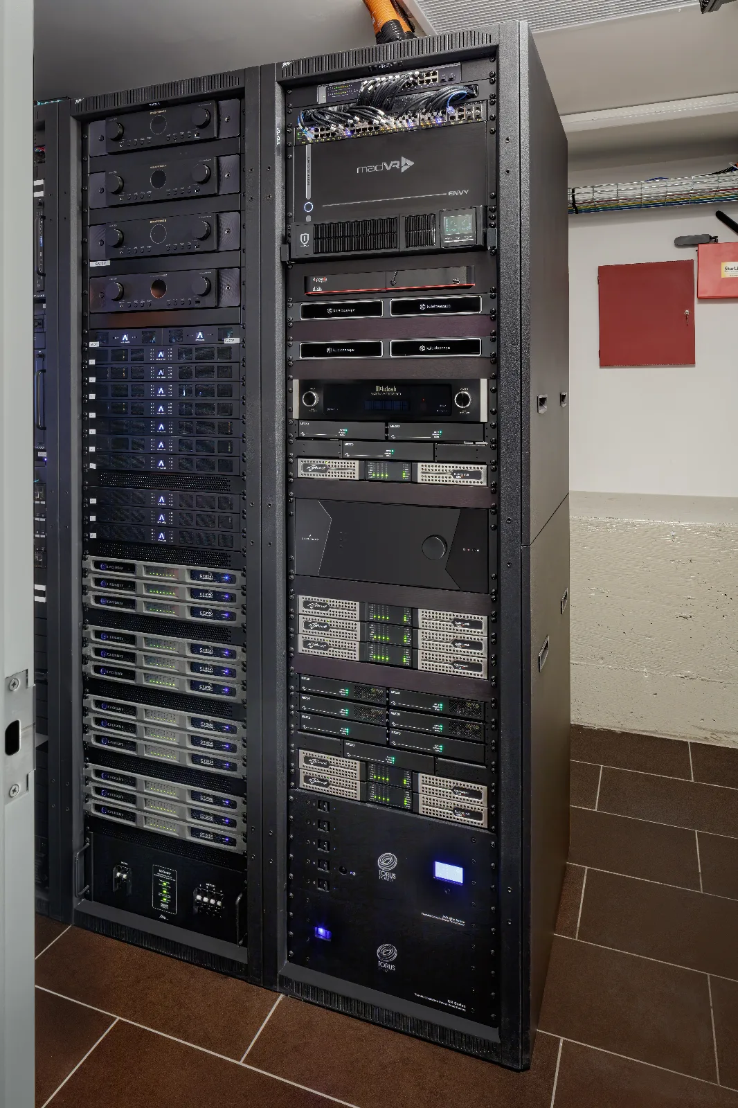 Two tall black server racks are densely packed with various electronic audio, video, and network equipment with blinking lights.