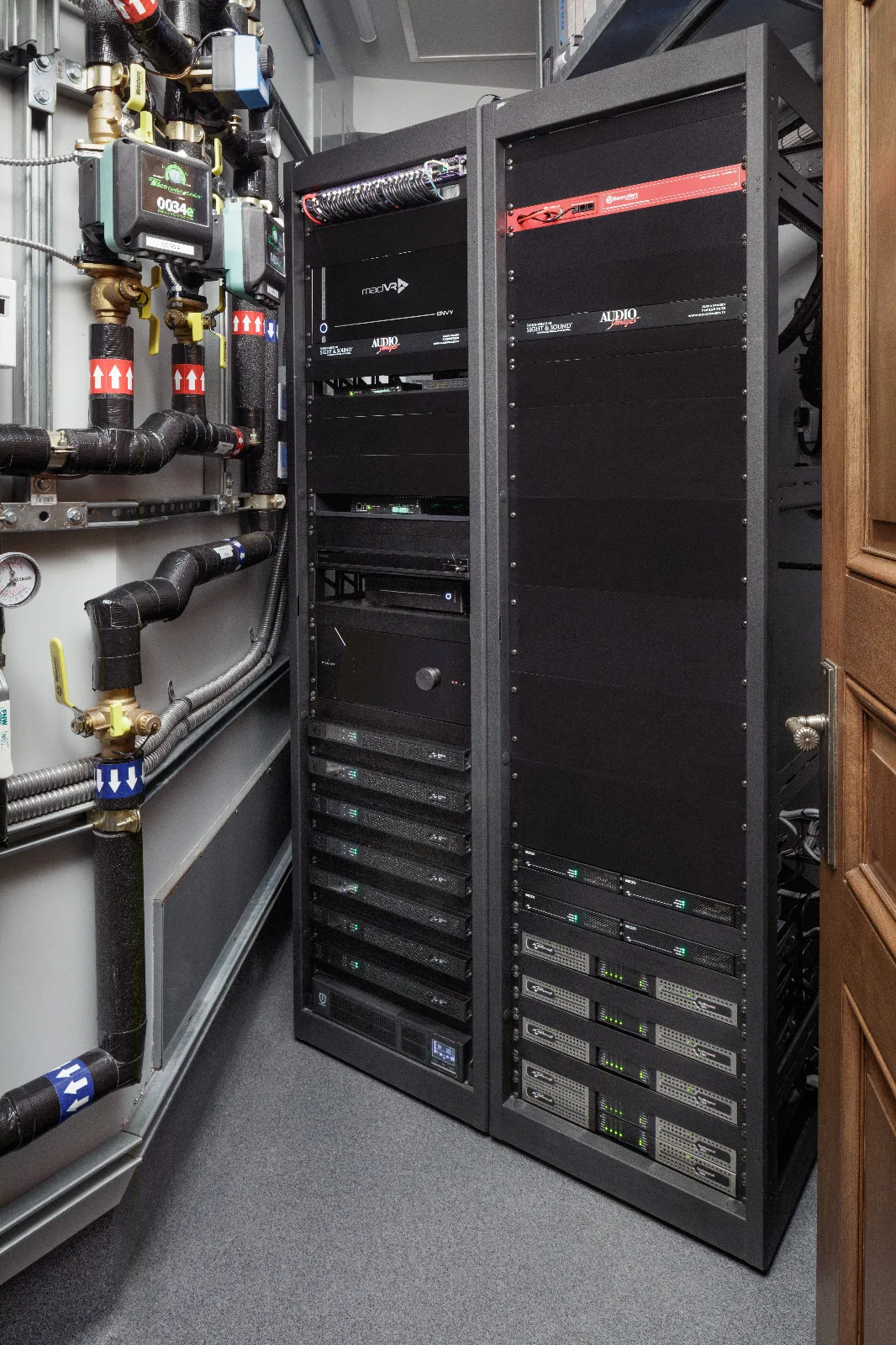 Two black server racks with audio and video equipment stand beside industrial pipes and a wooden door.